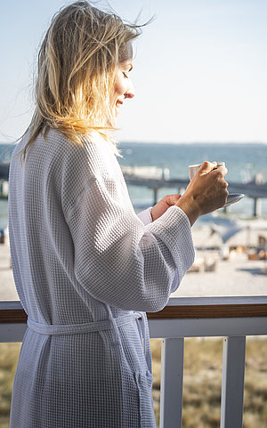 Frau steht im Bademantel auf dem Balkon und genießt ihren Kaffee mit Ausblick auf die Seebrücke Heiligenhafen und die Ostsee.
