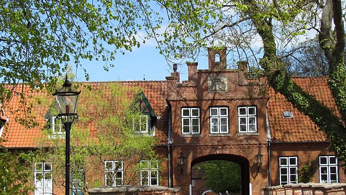 Ein Torhaus aus roten Steinen, mit Torbogen und weißen Fenstern. Dahin führt eine Brücke mit Kopfsteinpflaster.