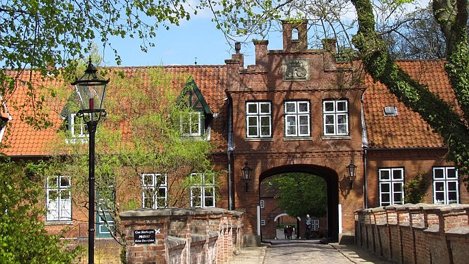 Ein Torhaus aus roten Steinen, mit Torbogen und weißen Fenstern. Dahin führt eine Brücke mit Kopfsteinpflaster.