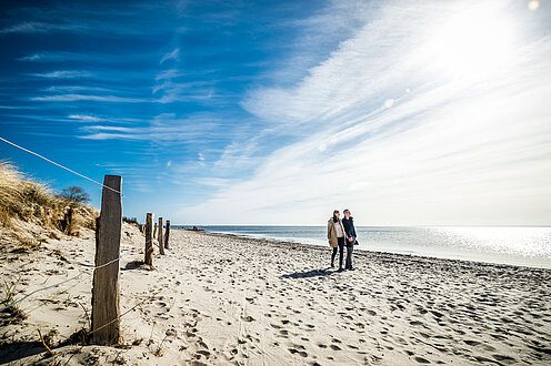 Ein Strand, klarer blauer Himmel, rechts im Bild Schleierwolken. Zwei Menschen gehen am Wasser spazieren. 