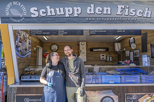 Ein Paar vor seiner Fischbude, sie schauen sich an und lächeln. 