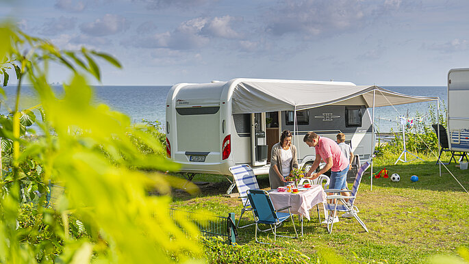 Zu sehen ist der Campingplatz in Surendorf bei Schwedeneck. Das Gras ist grün, der Himmel ist blau. Eine Familie hat ihr Wohnmobil in der Nähe der Steilküste abgestellt und ihren Tisch vor dem Wohnmobil aufgestellt. Von hier aus können sie den Ausblick auf die Ostsee genießen.