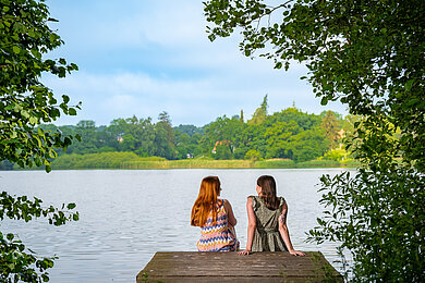 Zwei Frauen von hinten. Sie sitzen auf einem Steg am See und schauen auf den See, im Hintergrund bewaldete Wiesen. 