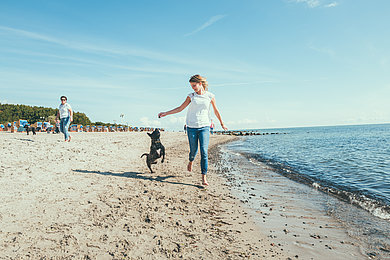 Eine Frau läuft mit einen Hund am Strand entlang. Beide haben sehr viel Spaß.