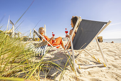 Zwei Frauen entspannen im Liegestuhl mit den Füßen im Sand in einer Strandbar an der Ostsee.
