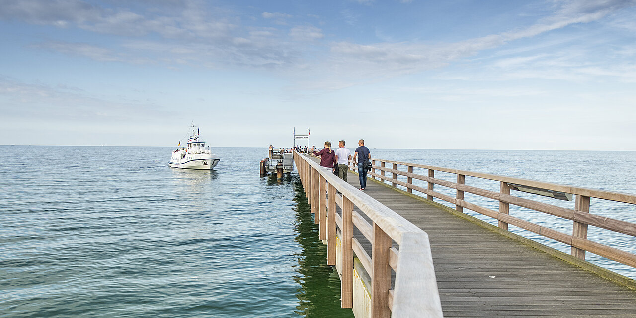 Schiff legt an einem sonnigen Tag an der Seebrücke Schönberg an. Viele Menschen stehen auf der Seebrücke. 
