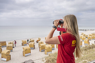 Blonde Frau mit rotem DLRG-T-Shirt schaut mit Fernglas aufs Meer.