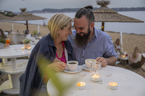 Ein Mann und eine Frau stecken die Köpfe zusammen und lachen in der Strandbar Tatort Hawaii. Sie trägt eine Decke um die Schultern, hinter ihnen geht die Sonne am Strand unter.
