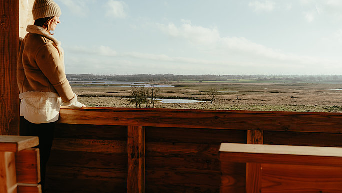 Eine FRau in Winterkleidung steht auf einem Aussichtsturm aus Holz und schaut auf die herbstlich-winterliche Landschaft der Hohwachter Bucht an der Ostsee.