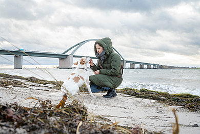 Eine Frau macht mit ihrem Hund Urlaub an der Ostsee SH. Sie sitzt entspannt in den Dünen am Strand von Fehmarn und genießt die herbstliche Atmosphäre. Sie streichelt liebevoll ihren Hund, der aufmerksam zu ihr aufblickt, während sie eine warme Tasse in der Hand hält. Die goldenen Gräser und der wolkenverhangene Himmel unterstreichen die ruhige, natürliche Stimmung. Im nächsten Moment entdecken sie bei einem Spaziergang die Fehmanrsundbrücke. Der Wind weht und die beiden lassen sich durchpusten.