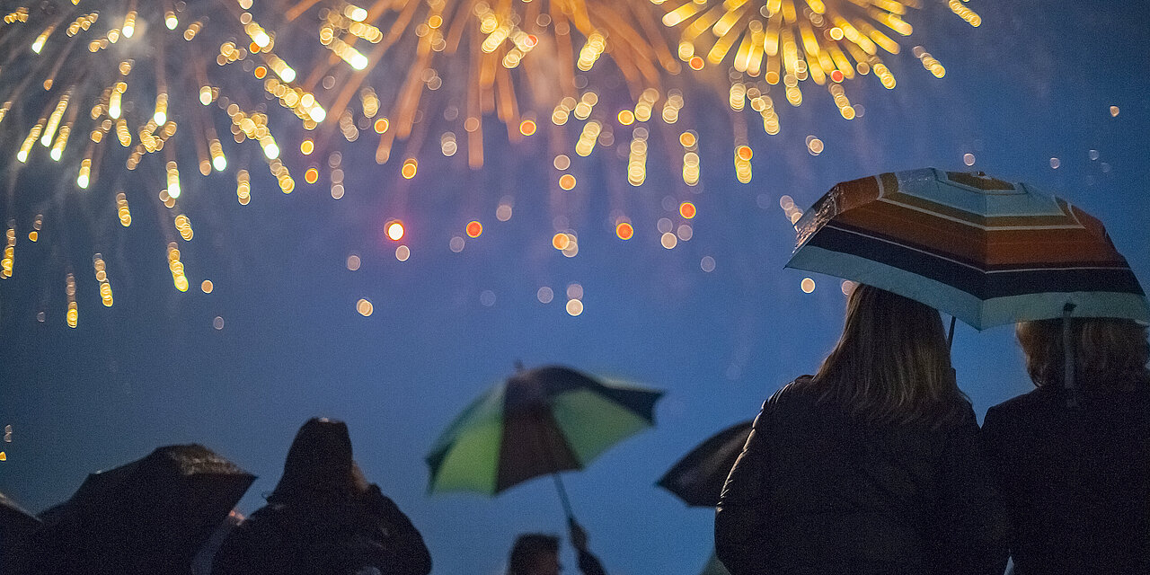 Viele Menschen stehen am Strand und beobachten das Feuerwerk, welches am Himmel passiert. Einige Menschen stehen mit Regenschirmen dort.