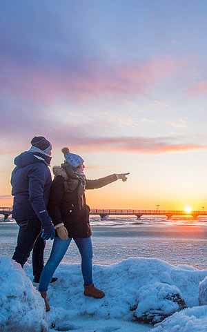 Ein Paar geht am Strand spazieren. Hinter ihnen ist die Ostsee zu sehen mit der Grömitzer Seebrücke. Am Strand liegen über all kleine weiße Schneehaufen. Das Paar hat dicke Winterjacken an, mit Schal und Mütze.