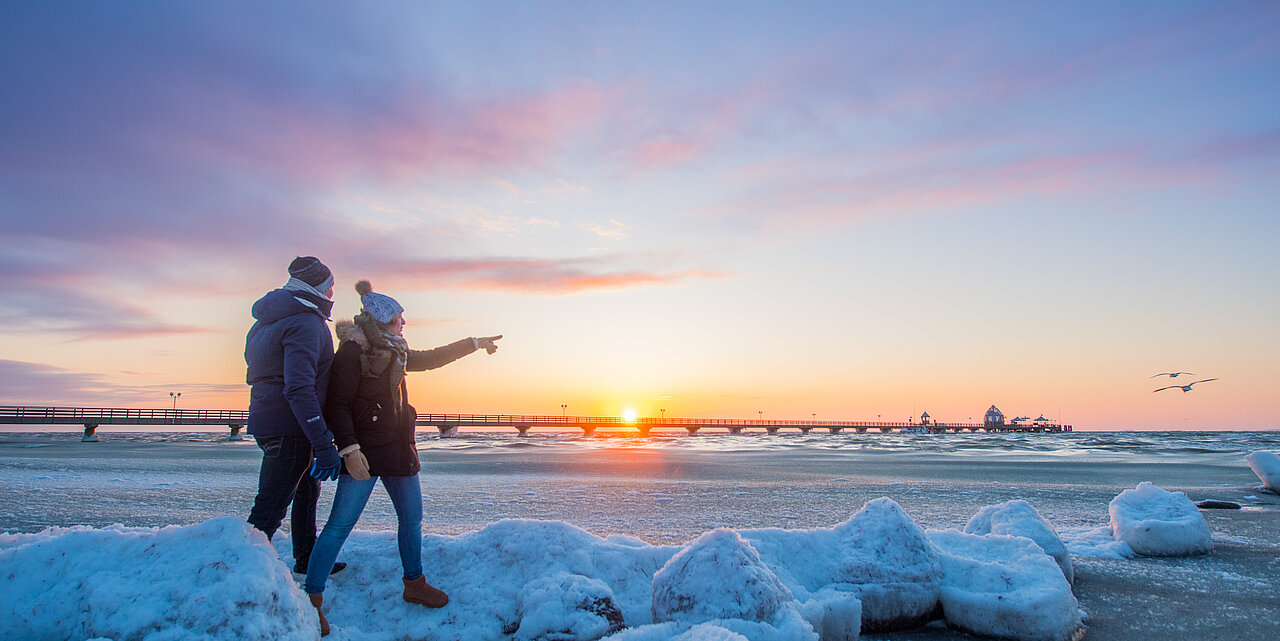 Ein Paar geht am Strand spazieren. Hinter ihnen ist die Ostsee zu sehen mit der Grömitzer Seebrücke. Am Strand liegen über all kleine weiße Schneehaufen. Das Paar hat dicke Winterjacken an, mit Schal und Mütze.