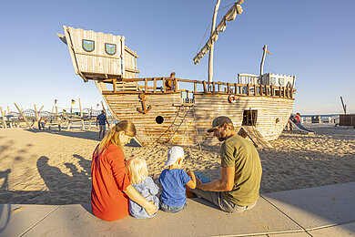 Familie sitzt auf den Stufen der Promenade. Sie schauen auf den Spielplatz am Strand. Dort steht ein großen Holzschiff (Spielgerät). Die Mama sitzt links, der Papa recht und dazwischen die beiden Kinder. Sie machen gerade eine kleine Pause und Essen ihre mitgebrachten Snacks.