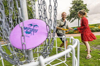 Paar spielt Discgolf im Kurpark von Eckernförde. Vorne im Bild sieht man den Basket mit einer Frisbee.