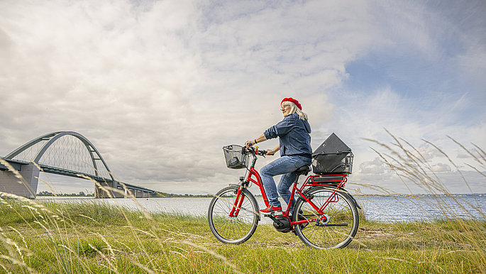 Eine Frau auf dem Rad, im Hintergrund das Meer und die Fehmarnsundbrücke. 