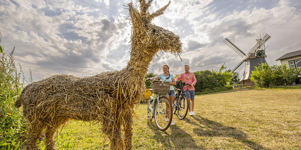 Paar radelt an den Probsteier Korntage die Radtour entlang den Strohfiguren. Auf dem Bild stehen sie gerade vor einem Reh, welches aus Stroh besteht.