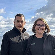 Ein Mann und eine Frau stehen eng beieinander, hinter ihnen das Meer.
