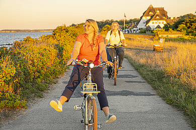 Radfahrer bei Abendlich. Eine Radfahrerin fährt auf den Betrachter zu, schaut links zum Meer und streckt die Beine vor und lässt sich rollen. Hinter ihr ein Radweg mit Feldern, im Hintergrund klein ein reetgedecktes Haus.