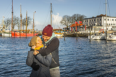 Paar umarmt sich. Im Hintergrund ist der Hafen von Laboe zu sehen. Es ist ein kalter sonniger Wintertag.