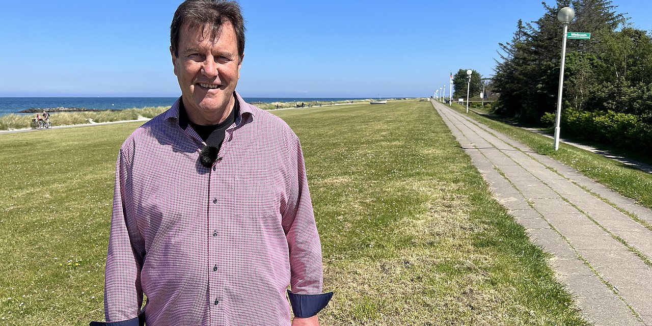 Ein Mann steh am Strand von Schönberg an der Ostsee. Im Hintergrund das Meer, neben ihm ein Radweg und eine Wiese. 
