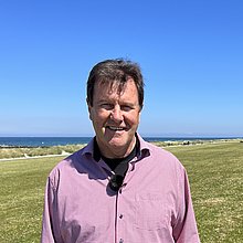 Ein Mann steh am Strand von Schönberg an der Ostsee. Im Hintergrund das Meer, neben ihm ein Radweg und eine Wiese. 