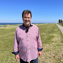 Ein Mann steh am Strand von Schönberg an der Ostsee. Im Hintergrund das Meer, neben ihm ein Radweg und eine Wiese. 