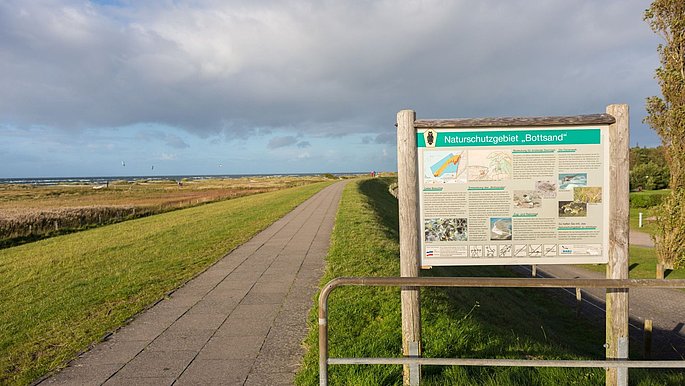 Zu sehen ist ein Wanderweg im Naturschutzgebiet Bottsand. Rechts neben dem Weg steht ein Informationsschild.