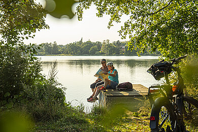 Paar macht während der Fahrradtour eine kleine Pause am See. Das Paar sitzt auf dem Steg und schaut in die Karte.