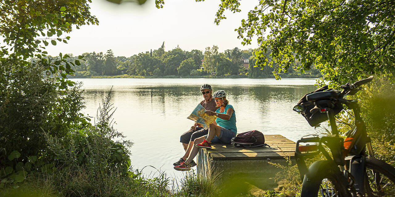 Paar macht während der Fahrradtour eine kleine Pause am See. Das Paar sitzt auf dem Steg und schaut in die Karte.