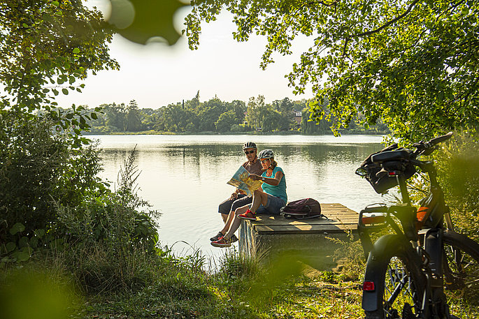 Paar macht während der Fahrradtour eine kleine Pause am See. Das Paar sitzt auf dem Steg und schaut in die Karte.