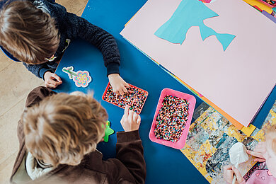 Ein Kindertisch von oben. Zu sehen sind Bastelsachen und Kinder, die etwas mit bunten Perlen basteln.