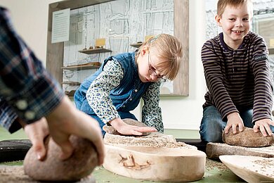 Zwei Kinder im Museum. Ein Mädchen beugt sich über einen hellen Stein und legt ihre Hand darauf. Ein Junge, daneben, lächelt und fasst ebenso auf ein Ausstellungsstück.
