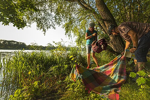 Zwei Personen machen eine kleine Picknickpause am See zwischen grünen Bäumen und Gräsern. Sie platzieren gerade ihre Picknickdecke.
