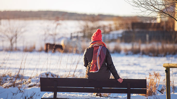 Eine Frau (dick gegen die eisige Kälte angezogen) sitzt auf der Rücklehne einer Bank und genießt die winterliche Landschaft mit Blick auf den See.