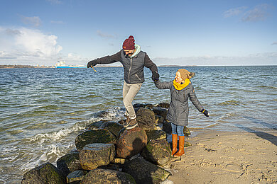 Paar geht am Strand in Wassernähe spazieren. Mann steht auf einem Stein, der am Strand im Wasser liegt. Es ist Herbst.