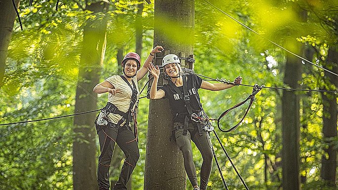 Zwei Frauen stehen auf einer Plattform, welche an einem Baumstamm befestigt ist. Beide tragen Kletterausrüstung und sind gesichert. Die Frau die Rechst steht winkt und die Frau die links steht, zeigt das Peace-Zeichen in die Kamera.
