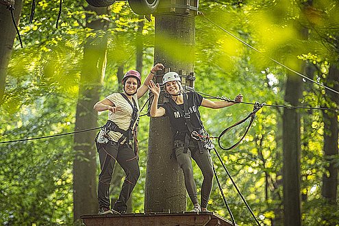 Zwei Frauen stehen auf einer Plattform, welche an einem Baumstamm befestigt ist. Beide tragen Kletterausrüstung und sind gesichert. Die Frau die Rechst steht winkt und die Frau die links steht, zeigt das Peace-Zeichen in die Kamera.
