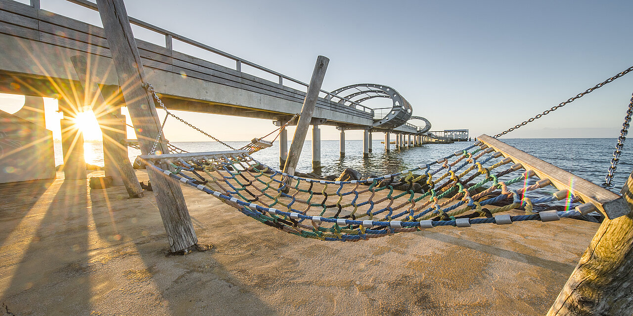 An Fuße der Seebrücke gibt es eine Plattform mit Hängematten. Eine ist direkt vorne im Bild zu sehen. Von da aus ist die Seebrücke zu sehen, wie sie aufs Wasser hinausfürht.