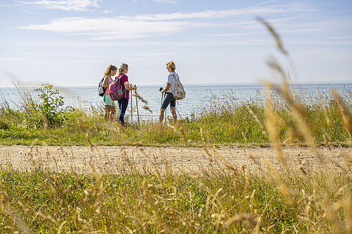 Von den Wiesen und einem Wanderweg geht der Blick Richtung Meer. Dort stehen drei Menschen, die auf einer Wanderung sind.