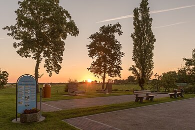 Ein Platz, auf dem Boukle gespielt werden kann. Die Sonne geht rot hinter ein paar grünen Bäumen unter.