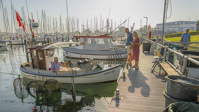 Paar geht am Fischereihafen von Strande spazieren. Links ist ein Fischerboot mit einem Fischer zu sehen.