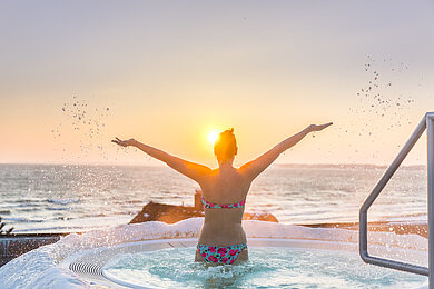 Frau steht im Whirlpool, der in der Außenanlage der Ostsee Therme ist, und streckt ihre lang zur Seite aus. Dabei blickt sie in die aufgehende Sonne über der Ostsee direkt vor ihr.