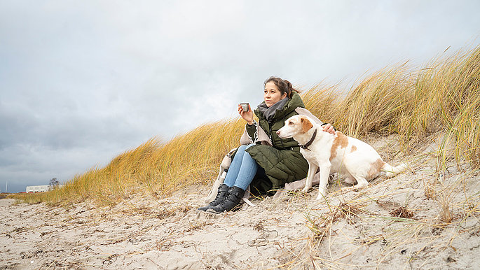 Eine Frau macht mit ihrem Hund Urlaub an der Ostsee SH. Sie sitzt entspannt in den Dünen am Strand und genießt die herbstliche Atmosphäre. Sie streichelt liebevoll ihren Hund, der aufmerksam zu ihr aufblickt.