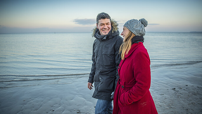 Ein Paar spaziert im Winter an der Ostsee entlang. Der Mann trägt eine blaue Winterjacke, die Frau eine rote Winterjacke.