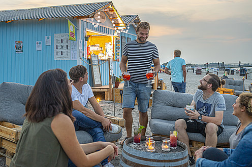 Viele Menschen sitzen in der Strandbar und trinken ein Getränk. Die Sonne geht langsam unter und die Lichterkette an der Strandbar im Hintergrund leuchtet.