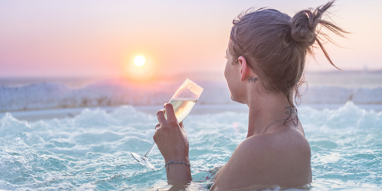 Eine Frau genießt im Whirlpool den Sonnenaufgang. In der Hand hält sie ein Sektglas, gefüllt mit Sekt. Sie schaut Richtung Sonnenaufgang. Der Himmel leuchtet in Pastellfarben von gelb über rötlich/rosa bis hellblau. Überall liegt weißer Schnee. . 