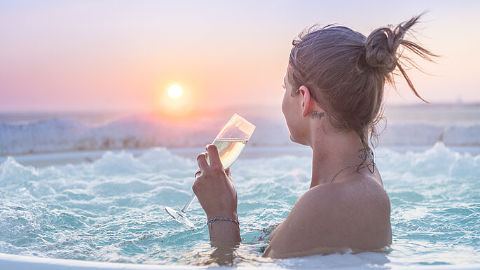 Eine Frau genießt im Whirlpool den Sonnenaufgang. In der Hand hält sie ein Sektglas, gefüllt mit Sekt. Sie schaut Richtung Sonnenaufgang. Der Himmel leuchtet in Pastellfarben von gelb über rötlich/rosa bis hellblau. Überall liegt weißer Schnee. . 