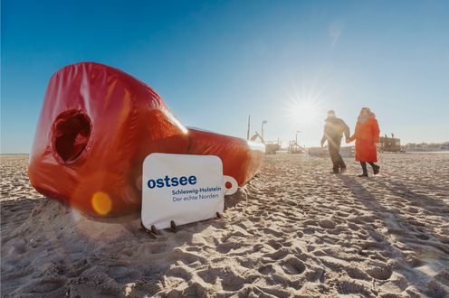 Paar geht auf die Riesenwärmflasche zu. Die Wärmflasche liegt am Ostsee-Strand.