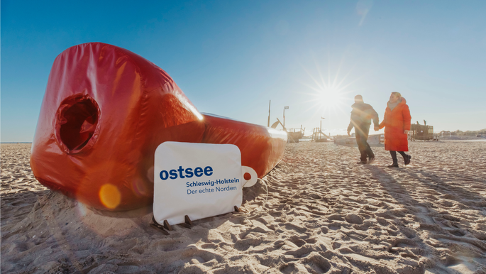 Paar geht auf die Riesenwärmflasche zu. Die Wärmflasche liegt am Ostsee-Strand.